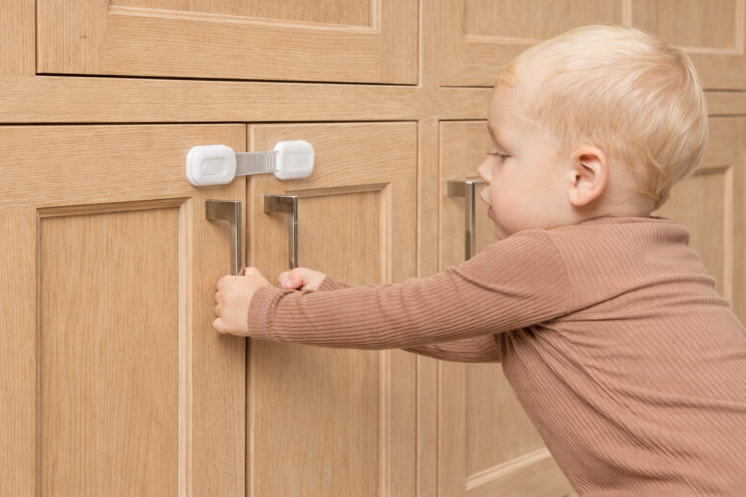 Toddler trying to open light wood kitchen cabinets secured with white child safety cabinet and drawer locks